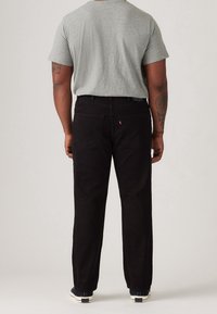 Pantalones de pana negros con corte recto, que tienen dos bolsillos traseros y una pequeña etiqueta de logo rojo cerca de la cintura. Combinados con una camiseta gris.