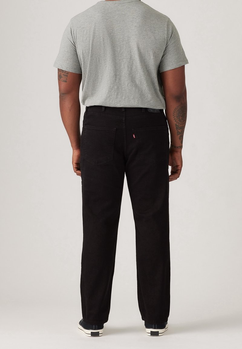 Pantalones de pana negros con corte recto, que tienen dos bolsillos traseros y una pequeña etiqueta de logo rojo cerca de la cintura. Combinados con una camiseta gris.