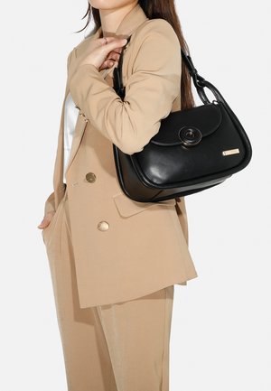 Borsa a tracolla in pelle nera con forma arrotondata, chiusura a grande bottone centrale e un delicato accento metallico. Indossata su un completo beige.