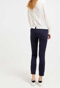 Blusa blanca con costura trasera con volantes combinada con pantalones azul marino oscuros; la modelo lleva zapatillas blancas con acentos de moño negro.