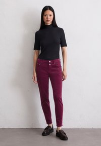 Schwarzes kurzärmliges Top mit Stehkragen, kombiniert mit bordeauxfarbenen Cordhosen, die eine schlanke Passform haben und zwei vordere Taschen mit Knopfdetails aufweisen.