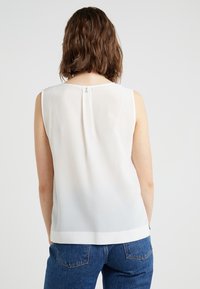 Beżowa bluzka bez rękawów z gładkiego, półprzezroczystego materiału, z małym guziczkiem z tyłu i prostym dołem, zestawiona z niebieskimi dżinsami.