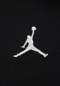 Tissu noir avec un logo brodé blanc représentant une figure sautant tenant un ballon de basket, mettant en valeur une texture lisse et des coutures détaillées.