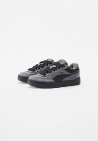 Un par de zapatillas Puma grises y negras con cordones negros y ojetes naranjas sobre un fondo blanco.
