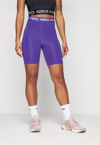 Shorts Nike Pro violets avec une taille haute, associés à des chaussures de sport rose clair et des chaussettes blanches. Tissu lisse avec une coupe ajustée.