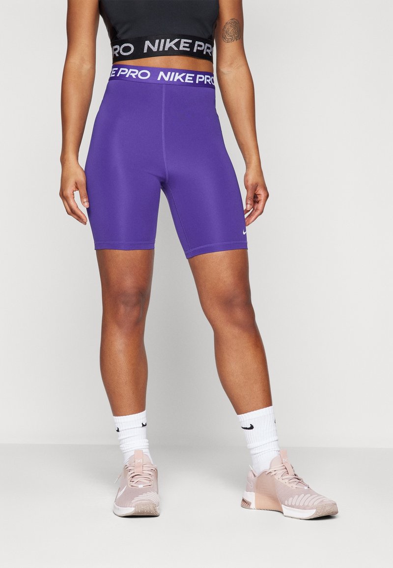 Shorts Nike Pro violets avec une taille haute, associés à des chaussures de sport rose clair et des chaussettes blanches. Tissu lisse avec une coupe ajustée.