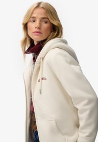 Femme blonde portant un sweat à capuche crème zippé sur une chemise à carreaux rouges, regardant sur le côté devant un fond blanc uni.