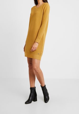 Woman wearing a mustard yellow ribbed long-sleeve sweater dress and black ankle boots standing against a plain white background.