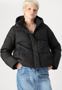 Černá péřová bunda s kapucí, s uzávěrem na zip, prošívaným designem a bočními kapsami. Nosená přes zelený top a riflovou sukni.