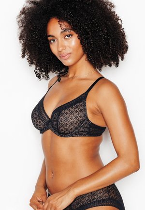 Donna con capelli ricci che indossa un reggiseno in pizzo nero e biancheria intima coordinata, in piedi davanti a uno sfondo bianco, sorridendo leggermente.