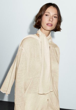 Maglione beige lavorato a maglia con maniche larghe, collo ampio e sciarpa fissata al collo; presenta una texture sottile e un dettaglio con bottone.
