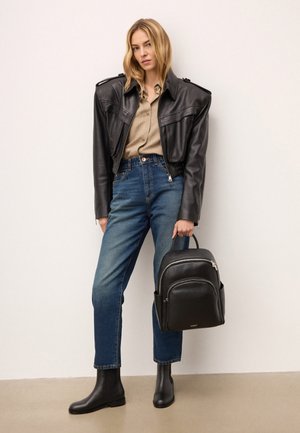 Sort læder rygsæk med rund form, lynlåsdetaljer og frontlomme. Modellen bærer en sort læderjakke, beige skjorte og blå jeans.