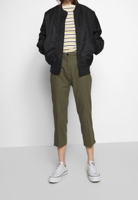 Svart bomberjacka, randig långärmad tröja med gula och marinblå ränder, olivgröna capribyxor och vita sneakers.