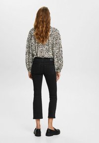 Mujer con cabello largo y ondulado, lleva una blusa de estampado de leopardo, jeans negros recortados y mocasines negros, de pie contra un fondo claro y sencillo.