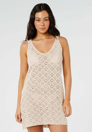Femme aux longs cheveux foncés portant une robe en crochet beige sans manches avec un col en V, se tenant devant un fond clair uni.
