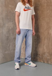 Valkoinen puuvillapaita, jossa on punainen ja musta Nike-logo, vaaleansiniset farkut ja valkoiset lenkkarit, joissa on vihreitä ja oransseja yksityiskohtia. Yksinkertainen tausta.