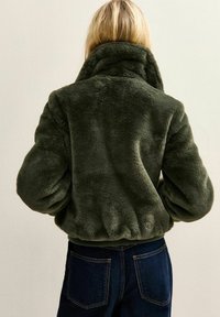 Persona rubia que lleva una chaqueta de piel sintética verde oscuro con cuello alto y vaqueros azul oscuro, de espaldas.