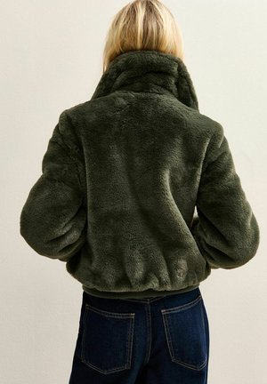 Persona rubia que lleva una chaqueta de piel sintética verde oscuro con cuello alto y vaqueros azul oscuro, de espaldas.