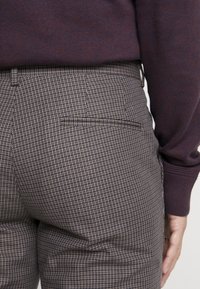 Kostkované kalhoty v hnědé a krémové látce, se jednou zadní kapsou a upraveným střihem. Spárovány s tmavě vínovým svetrem.