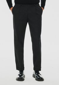 Czarne, dopasowane spodnie o gładkiej fakturze, z przodu z zaszewkami, bocznymi kieszeniami i podwiniętym nogawkami, w połączeniu z błyszczącymi czarnymi butami.