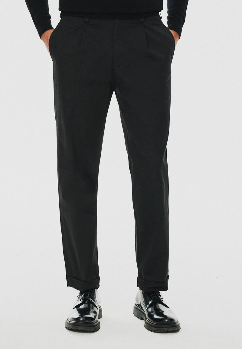 Czarne, dopasowane spodnie o gładkiej fakturze, z przodu z zaszewkami, bocznymi kieszeniami i podwiniętym nogawkami, w połączeniu z błyszczącymi czarnymi butami.