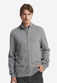 Grå zip-up cardigan i mjukt stickat tyg, med hög krage, ribbade muddar och fåll, i en rak silhuett och subtil textur.