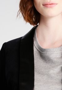 Černé sako s lesklými saténovými klopami přes šedou mikinu s kulatým výstřihem a měkkou texturou, zobrazena od límce vzhůru.