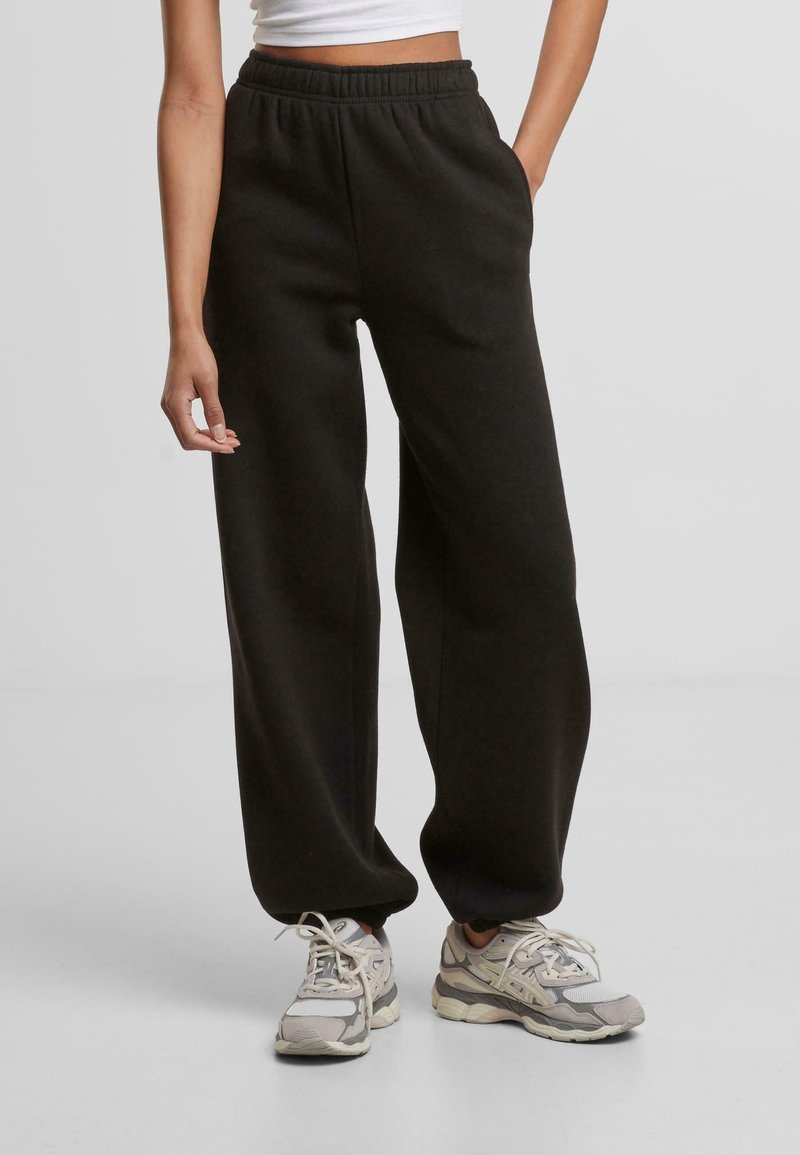 Femme portant un pantalon de jogging noir ample avec une taille élastique et des baskets de sport grises et blanches, se tenant avec une main dans la poche.