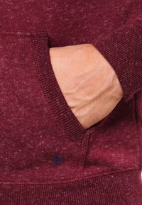 Bordeauxrode trui van zacht, gestructureerd stof, met geribbelde manchetten en een voorvak. Voorzien van een klein blauw logo op het vak.