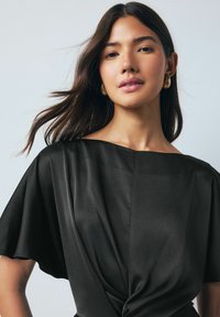 Fekete, textúrált blúz széles, hajónyakú és rövid, laza ujjakkal. Csavart részlettel rendelkezik a deréknál, amely laza szabást biztosít.