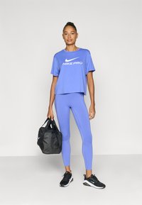 Μπλε κοντομάνικη μπλούζα Nike Pro με λευκό λογότυπο, σετ με αντίστοιχο μπλε κολάν. Μαύρα αθλητικά παπούτσια και μαύρη τσάντα duffel με λογότυπο.