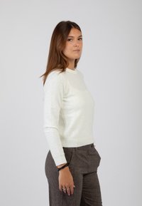 Maglione in maglia color crema chiara con design aderente, delicato motivo testurizzato e dettagli a coste sull'orlo, indossato con pantaloni marroni sartoriali.