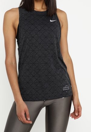 Frau trägt ein ärmelloses dunkelgraues gesteppte Nike-Tanktop und glänzende graue Leggings und steht vor einem weißen Hintergrund.