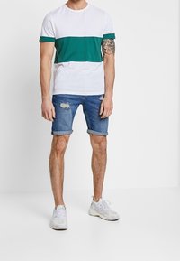 Miestenské tričko s bielym telom a zeleným horizontálnym pruhom, v kombinácii s roztrhnutými denimovými šortkami a bielymi teniskami.