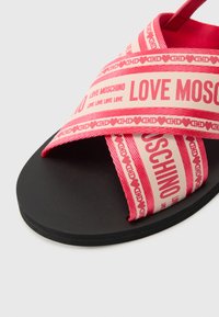 Roza sandali s križnim trakami in tiskanim besedilom "LOVE MOSCHINO" na črnem ravnem podplatu. Gladek tekstilni zgornji del.
