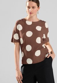 Haut marron à manches courtes avec de grands cercles crème, doté d'un col rond et d'une coupe décontractée, associé à un pantalon noir contrasté.