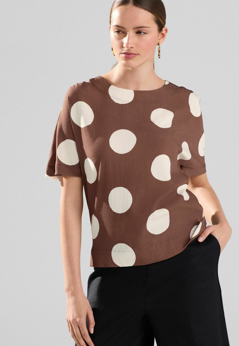 Haut marron à manches courtes avec de grands cercles crème, doté d'un col rond et d'une coupe décontractée, associé à un pantalon noir contrasté.