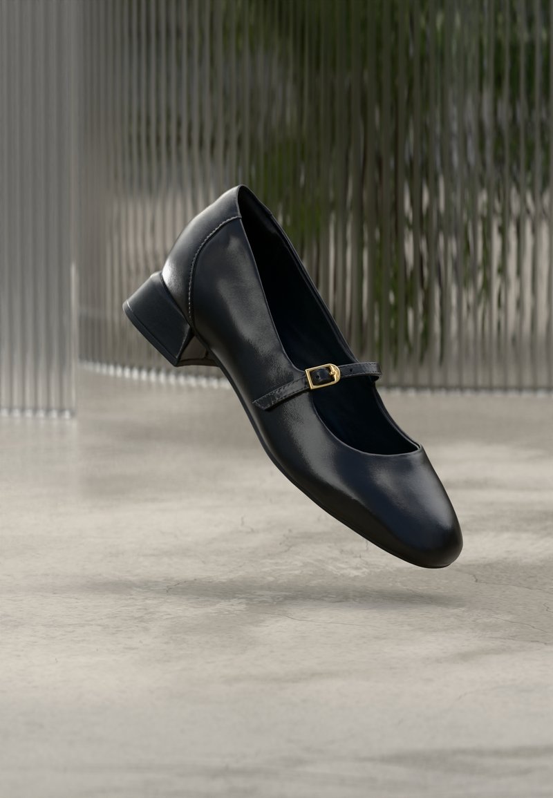 Chaussure Mary Jane en cuir noir avec un petit talon carré et une boucle dorée flottant au-dessus d'un sol en béton, avec un arrière-plan texturé translucide.