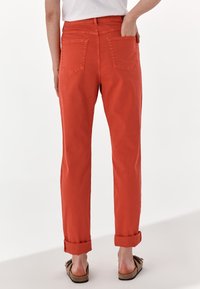 Red cotton pants with a straight leg design, featuring rolled cuffs and two back pockets. Worn with sandals. Background is plain white.