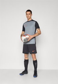 Grå og svart atletisk trøye med røde detaljer, svarte shorts, svarte fotballsko, og leggbeskyttere holdt av en person med tatoveringer, som holder en mønstret fotball.