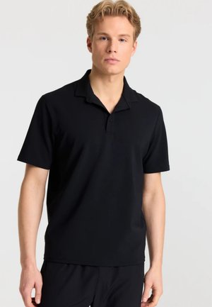 Ung mand med lyst hår iført en ensfarvet sort kortærmet polo-shirt mod en hvid baggrund.