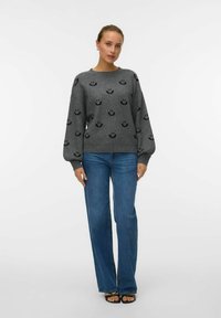 Szary sweter z wzorem uśmiechniętej buzi, szerokimi rękawami i ściągaczami, w połączeniu z niebieskimi dzwonami i czarnymi sandałami.