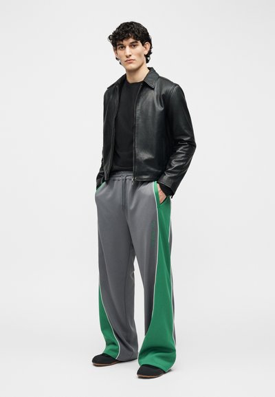 Νέος άνδρας που στέκεται με τα χέρια στις τσέπες, φοράει μαύρο δερμάτινο μπουφάν, μαύρο πουκάμισο και φαρδιά γκρι παντελόνια με πράσινες πλαϊνές λωρίδες και μαύρα παπούτσια.