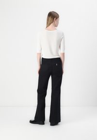 Pantalons évasés noirs avec une fermeture éclair et un bouton, dotés de deux poches arrière et d'un petit logo métallique. Portés avec un haut blanc à manches courtes.