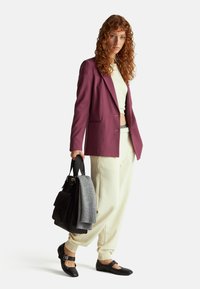 Bordowy żakiet na jasnej bluzce, kremowe dresy, czarne buty z klamrami, trzymający czarną torbę i szary sweter, długie kręcone włosy.