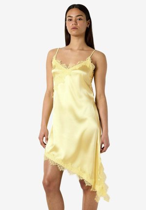 Kvinde iført en ærmeløs gul satin slip kjole med blonder og asymmetrisk nederdel, stående foran en ensfarvet hvid baggrund.