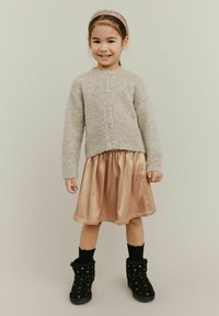 Harmaa neulecardigan napilla, satiini ruusukultainen hame, mustat saappaat hopeaisilla yksityiskohdilla, mustat sukat, lapsi poseeraa neutraalin taustan edessä.