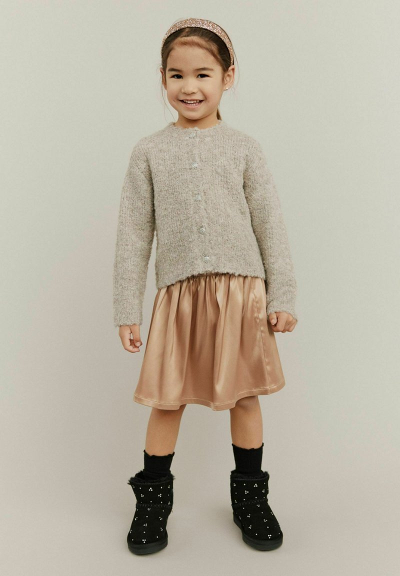 Harmaa neulecardigan napilla, satiini ruusukultainen hame, mustat saappaat hopeaisilla yksityiskohdilla, mustat sukat, lapsi poseeraa neutraalin taustan edessä.