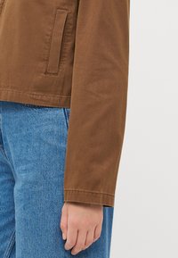 Jachetă din bumbac maro cu o textură netedă, având o croială dreaptă și un buzunar lateral, asortată cu blugi din denim de culoare albastru deschis.