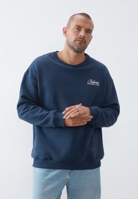 Felpa blu navy con colletto rotondo, polsini e orlo a costine. Presenta un logo ricamato bianco con testo sul petto. Texture del tessuto morbida.